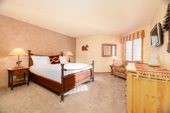 A cozy bedroom featuring a king-size bed, rustic decor, a television, and a window with blinds, creating a warm and inviting atmosphere at the Snow Park Chalet.