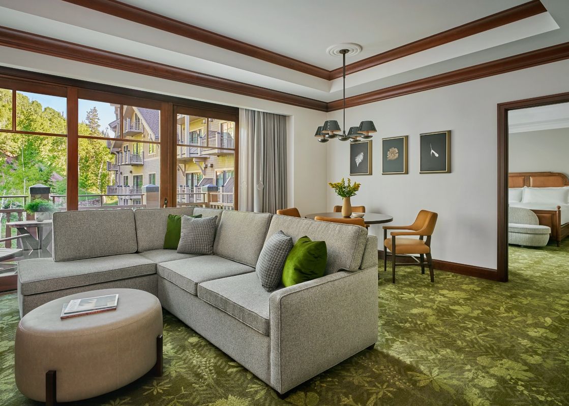 A spacious living area features a large L-shaped sofa, a round ottoman, and a dining table with chairs, all set on a green patterned carpet, with a large window offering a view of lush trees and buildings.