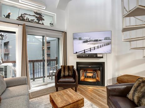 A cozy living room featuring a flat-screen TV above a fireplace, a grey sofa, two brown leather armchairs, a wooden coffee table, and a balcony with a view of a winter landscape.
