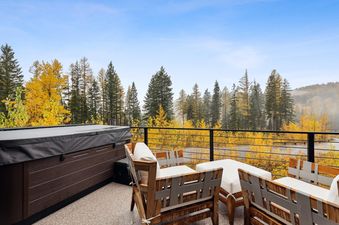 An outdoor patio at Snowghost Chalet features a hot tub and comfortable seating, overlooking a stunning autumn forest with bright yellow trees under a clear blue sky.