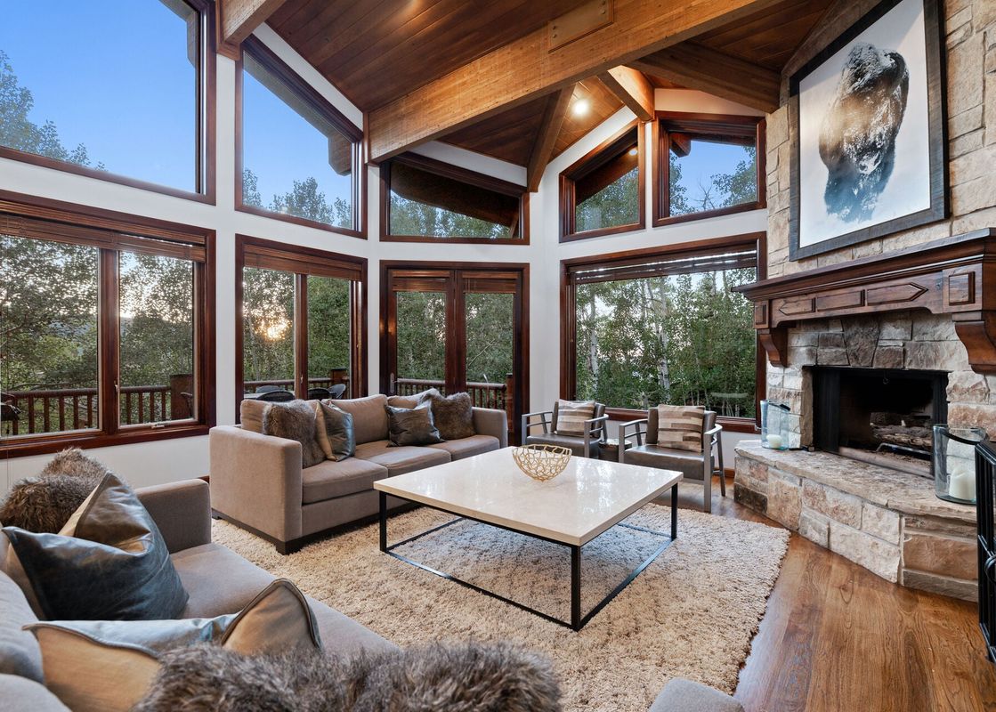 A bright, cozy living room in the Deer Valley Double Eagle cabin featuring vaulted wooden ceilings, large wrap-around windows with forest views, and a stone fireplace.