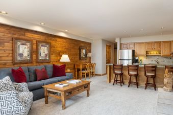 A wide view of the open-concept living room and kitchen area at Montaneros 314, featuring a gray sofa, patterned armchair, wooden coffee table, wood-paneled wall, and a kitchen with wooden cabinets and a breakfast bar.