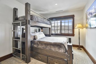 A cozy bunk room at Arrowleaf Lodge 311 with a custom dark wood bunk bed, plush grey bedding, a floor lamp, and a large window overlooking a snowy mountain landscape.
