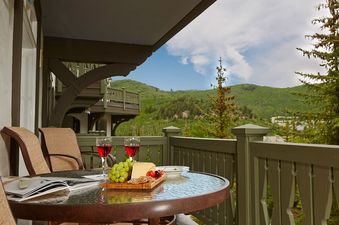 A private balcony at Chateau Beaver Creek Residence 309 featuring a round glass table set with wine, grapes, cheese, and crackers, offering views of green mountains under a blue sky.