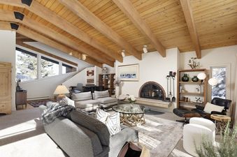 A spacious living room at Lupine Estate with a vaulted wooden beam ceiling, a large gray sofa, a glass coffee table, and a brick fireplace, offering a cozy and rustic atmosphere with snowy outdoor views.