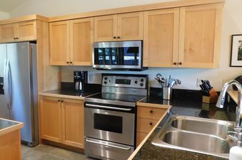 A modern kitchen with light wood cabinets, dark granite countertops, and stainless steel appliances, including a refrigerator, oven, microwave, and double-basin sink.