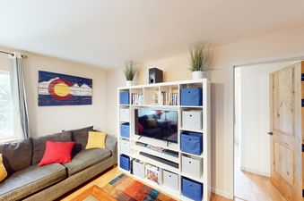 Living room at Beaver Creek Basecamp with a couch, white entertainment center with a TV and shelving, and an open door leading to another room.