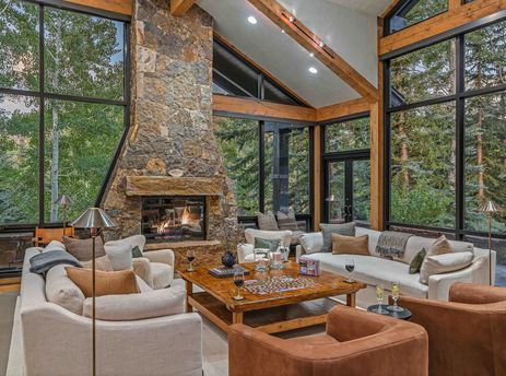 A living room with a fireplace and large windows overlooking a forest, a couch, two armchairs, and a coffee table with a puzzle on it