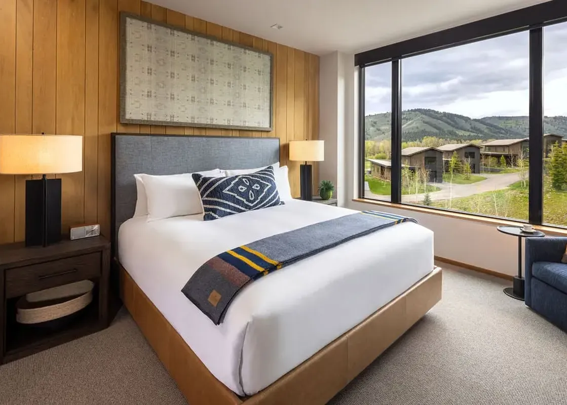 A modern guest room with a king-size bed, featuring a gray headboard and white linens, a striped throw blanket, and a large window overlooking a scenic mountain landscape with cabins.