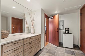 The entryway at Lowell 401A features a full-length mirror, a wooden dresser with a granite top, decorative vases, an open wooden door, a mini-fridge, and a coffee maker.