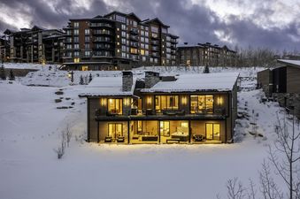 A luxurious modern chalet covered in snow at dusk with glowing interior lights.