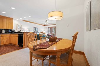 A cozy dining area with a wooden oval table and four chairs, leading into a kitchen and living room with a view of the snowy outdoors.