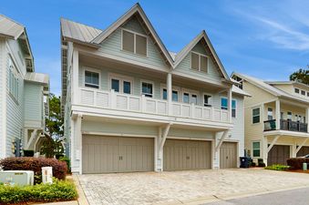 A modern coastal home with light blue siding, a metal roof, a second-story balcony, and multiple garage doors.