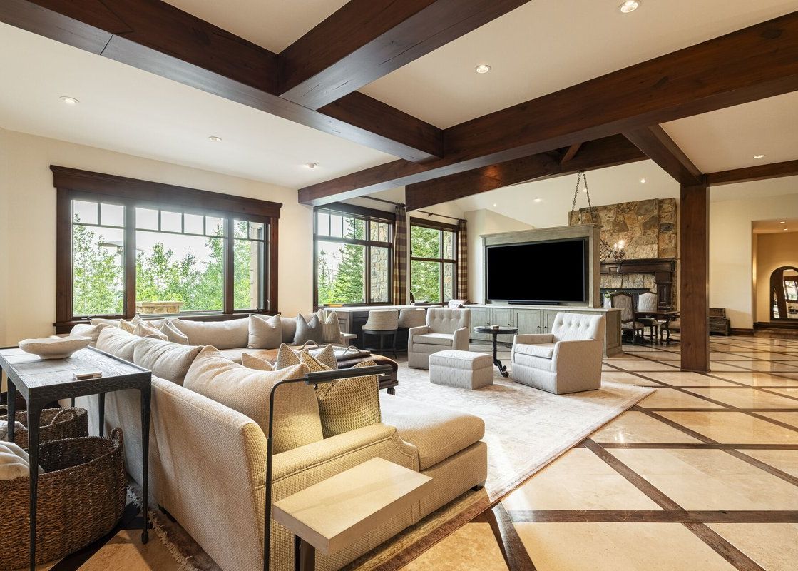 An expansive luxury living room featuring dark wooden ceiling beams, cozy seating arrangements, and large windows with scenic forest views.