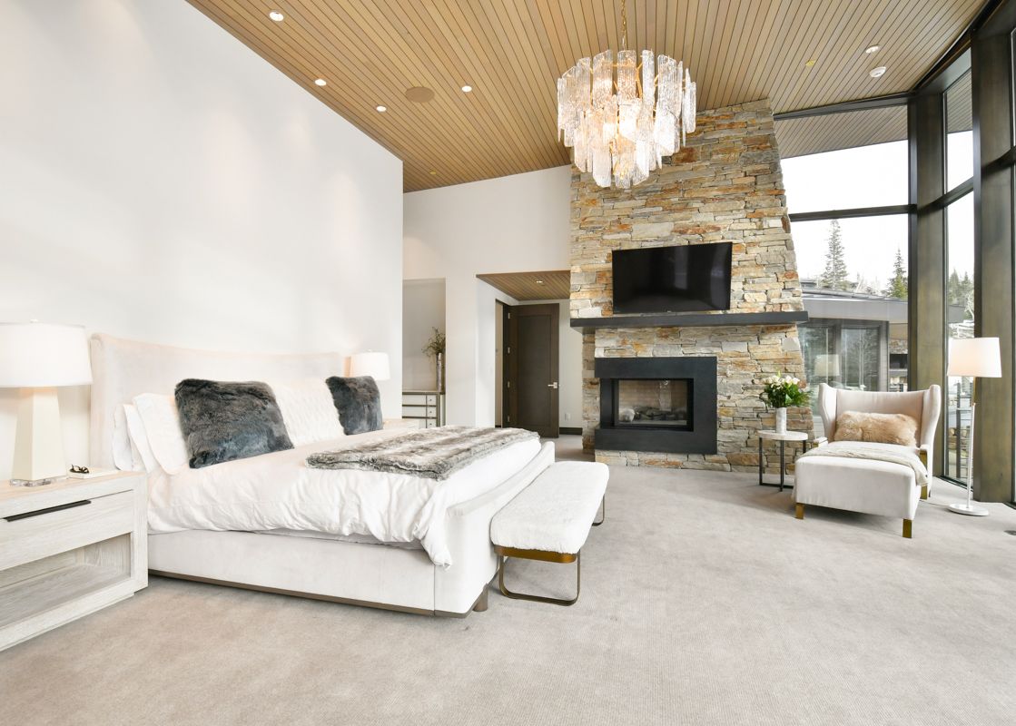 The primary bedroom features a king-sized bed, a stone fireplace with a mounted television, and a cozy armchair, all under a wooden slat ceiling.