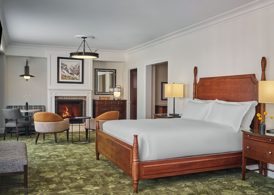 A spacious Montage Deer Valley Accessible Resort View Junior Suite with a large wooden bed, white bedding, a cozy fireplace, a small dining area, and comfortable seating, all bathed in warm lighting.