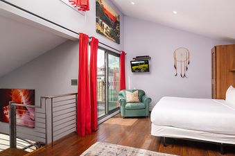 A cozy bedroom with a queen-sized bed, a green armchair, and a wall-mounted TV, featuring red curtains and artwork.