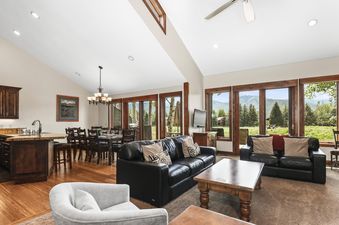A bright and spacious open-concept living and dining area with black leather sofas, a wooden coffee table, a dining table, and large windows offering mountain views.