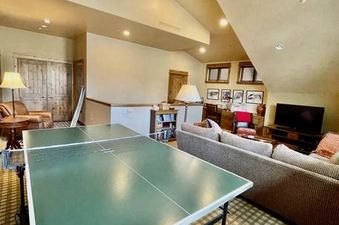 A spacious games room features a green ping pong table under a wooden ceiling fan, with comfortable couches and a TV visible in the background, making it an inviting space for recreation and relaxation.