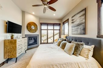 A modern master bedroom at Blackstone Residence 1-B features a large bed with decorative pillows, a wall-mounted TV, a fireplace, and scenic window views.