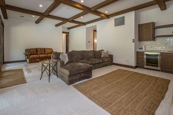 The image shows the living room of Stony Creek Chalet featuring two large couches, a coffee table, and an entertainment setup. A bar area with a wine cooler is visible to the right. The room is decorated with wood beams on the ceiling, two rugs, and light