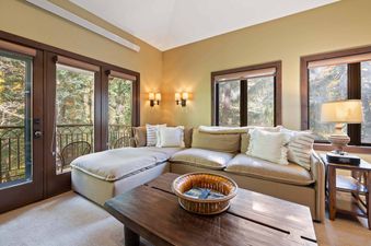 A cozy lounge area in The Charter C2105 featuring an L-shaped sofa, a wooden coffee table, and French doors leading to a balcony with a forest view.
