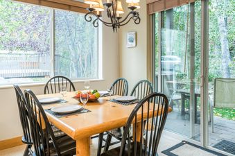 A dining room table with six chairs and a window overlooking a forest, with a sliding door opening to a patio.