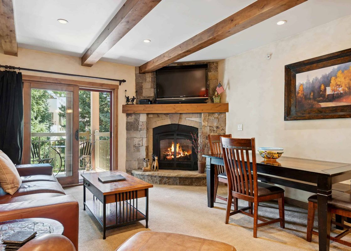 A cozy living room with a fireplace, a leather couch, a coffee table, a dining table with chairs, and a view of trees from the windows.
