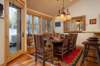 Warmly lit dining table set for six with rustic log chairs over a vibrant red rug.