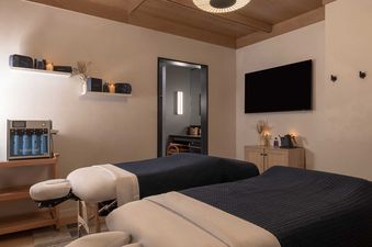 A modern and serene spa treatment room features two massage beds, a HydraFacial machine, and a wall-mounted television, creating a luxurious and relaxing atmosphere.