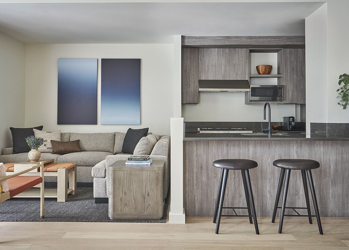 Pendry Two Bedroom Resort King Residences features a living area with a sectional sofa, two armchairs, a coffee table, two side tables, a rug, and artwork on the wall. There is a small kitchen with a microwave, stovetop, sink, and cabinets.
