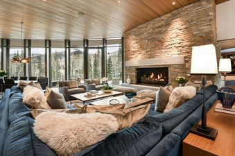 A luxurious living room at Iron Mountain Chalet featuring a large blue sectional sofa, a stone fireplace, and floor-to-ceiling windows overlooking a snowy forest.