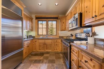 A rustic-chic kitchen with light wood cabinets, stainless steel appliances, stone countertops, and a window overlooking a mountainous, tree-filled landscape.
