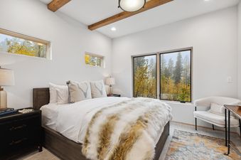 A cozy bedroom at Snowghost Chalet features a bed with white linens and a faux fur throw, rustic nightstands, and large windows with autumn forest views.
