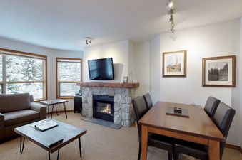 A cozy living space in Aspens on Blackcomb Unit 443 with a stone fireplace and a dining area, featuring large windows with a snowy winter view.