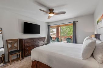 A master bedroom with a king-size bed, a large flat-screen TV above a wooden dresser, a ceiling fan, and a private balcony overlooking a scenic landscape with trees and snow.
