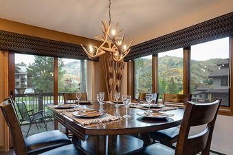 The dining room at the Ranch at Steamboat RA401, featuring a large wooden table set for eight, an antler chandelier, and expansive windows with views of mountains and autumn trees.