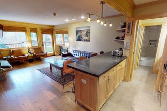 A cozy living room with a fireplace, comfortable seating, and a large window overlooking a wooded area. There is a kitchen island in the background.