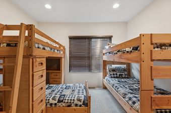 A cozy bedroom in Slopeside 117 featuring two wooden bunk beds with plaid and bear-themed bedding, a window with closed blinds, and carpeted flooring.