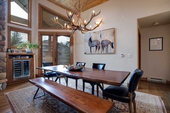 A rustic dining room with a large wooden table, black leather chairs, and a wooden bench, illuminated by an antler chandelier, with a wine cooler built into a wooden cabinet.