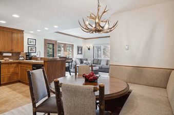 A rustic-chic dining area with a round wooden table, log chairs, and an antler chandelier, flowing into a modern kitchen and a cozy living room with large windows.