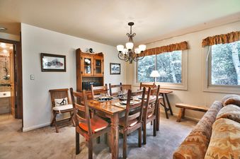 A cozy dining area at Snow Flower 148 features a wooden table set for six, illuminated by a chandelier and natural light from large windows offering a tree view. The room is adorned with framed art and a rustic hutch.