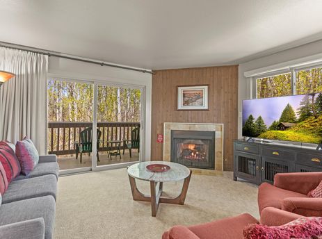 The inviting living room at Snow Flower 140 Park City features a cozy fireplace, comfortable seating, and a large flat-screen TV, all opening onto a balcony with a scenic wooded view.