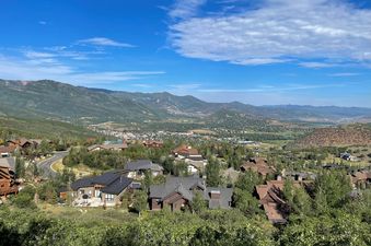 A panoramic mountain vista showcasing the Solamere Home nestled among lush green trees, with a charming town visible in the valley below, all under a clear blue sky.