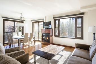 A spacious living room with a fireplace and a large window with a view of a snowy forest. The room is furnished with a couch, coffee table, and a dining table. The fireplace is lit and the room is bathed in sunlight.