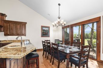 A spacious dining area with a long wooden table set for a meal, a multi-bulb chandelier, and an adjacent kitchen island with granite countertops and bar stools in Fairway Village 2919.