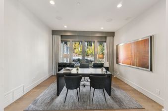 A modern executive office featuring a large dark wood desk with two grey chairs, set on a grey rug, with expansive windows overlooking autumn trees.