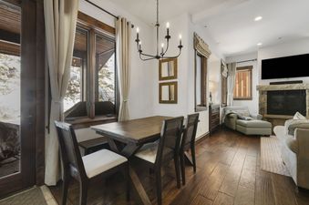 A dining table with chairs and a view of a snowy landscape through large windows, with a living area featuring a fireplace in the background at Juniper Landing 108.