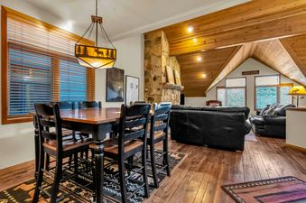 The dining room of Ski House on Lowell has a wood table, four chairs, and a rug. There are three windows with blinds, as well as a chandelier with moose silhouettes. The living room features a fireplace, couches, and a coffee table.