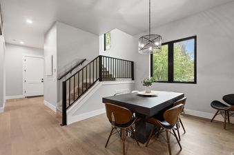 A modern dining area with a dark wood table and chairs, illuminated by a contemporary pendant light, and a staircase with a black railing, all set against a backdrop of light grey walls and large windows with an exterior view.